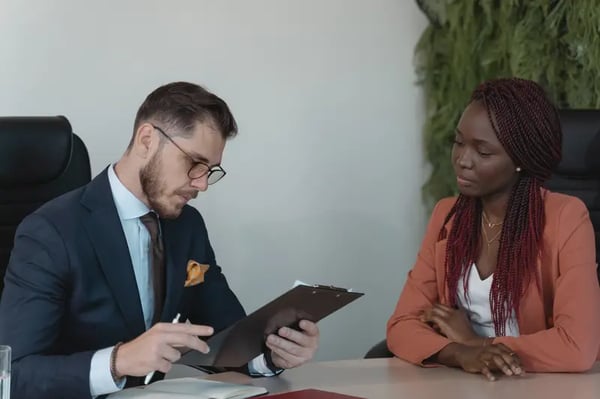 Two employees sitting at desk with one employee holding clipboard and another employee listening