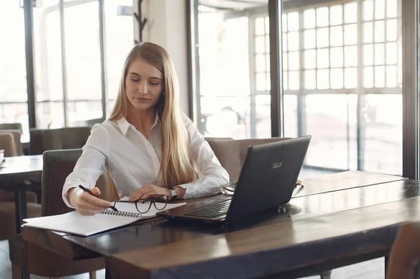 Woman sitting at desk writing in a book with laptop in front of her 