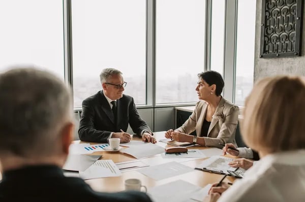 Two employees sitting at office desk talking with each other and two other employees blurred 