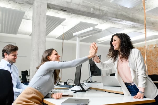 Two female employees highfiving across office table and smiling