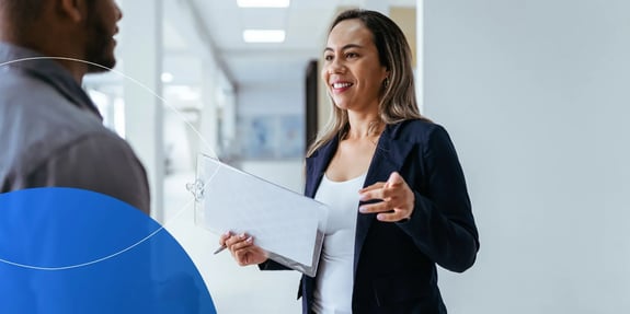 Woman standing in office setting holding clipboard and talking to person in front of her 