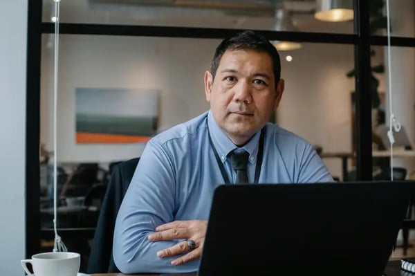 Male employee sitting on desk with laptop and looking into camera