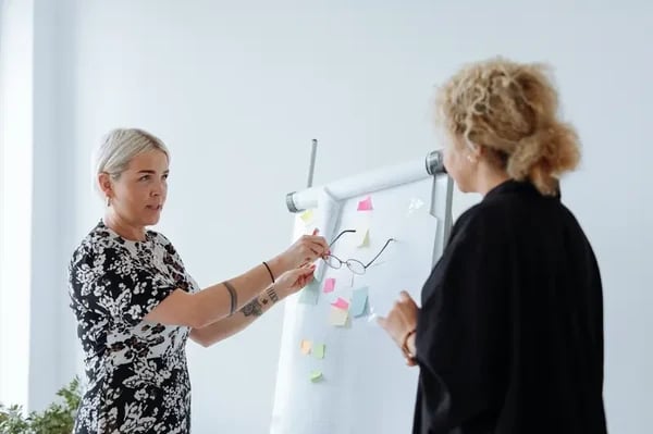 Two women standing in front of white board discussing post-it notes