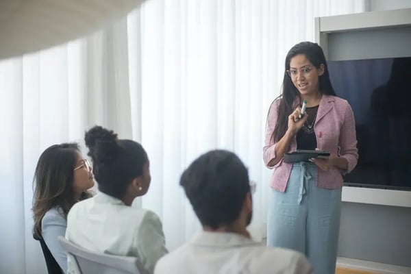 Woman holding tablet and market talking to three employees siting in front of her 