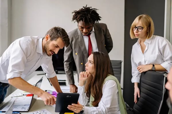 Four employees gathered around desk looking into tablet that a woman is holding 