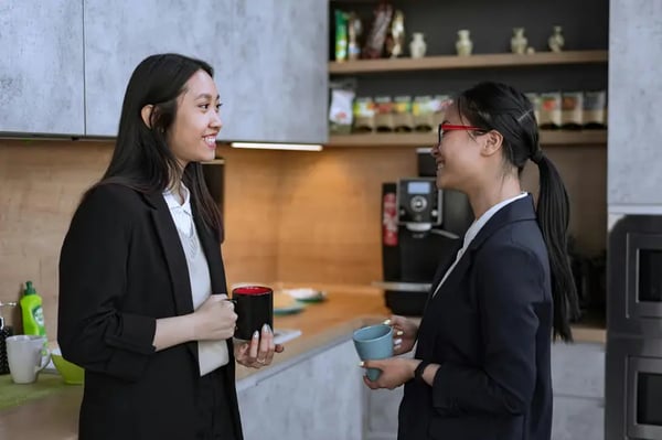 Two female employees holding coffee mug and smiling at each other in office kitchen 