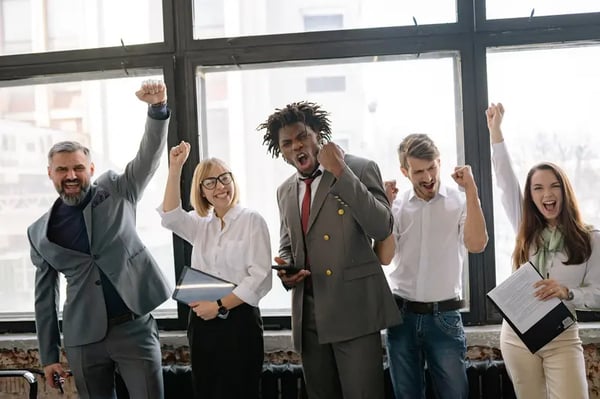 Five employees standing infront of office window with fists in the air showing excitement