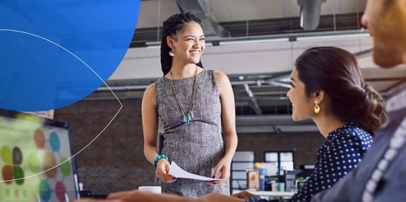 Woman standing and smiling at other female employees holding paper in hand 