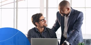 Männlicher Angestellter, der mit einem Laptop am Schreibtisch sitzt und zu einem anderen männlichen Angestellten aufschaut und lächelt