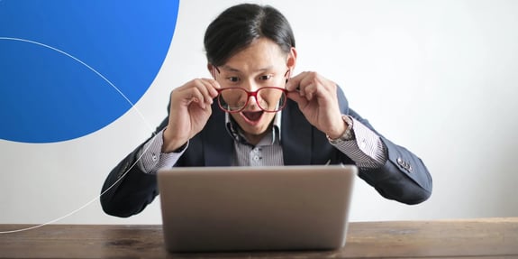 Man holding the glasses he is wearing away from his face and looking into the laptop with a shocked expression