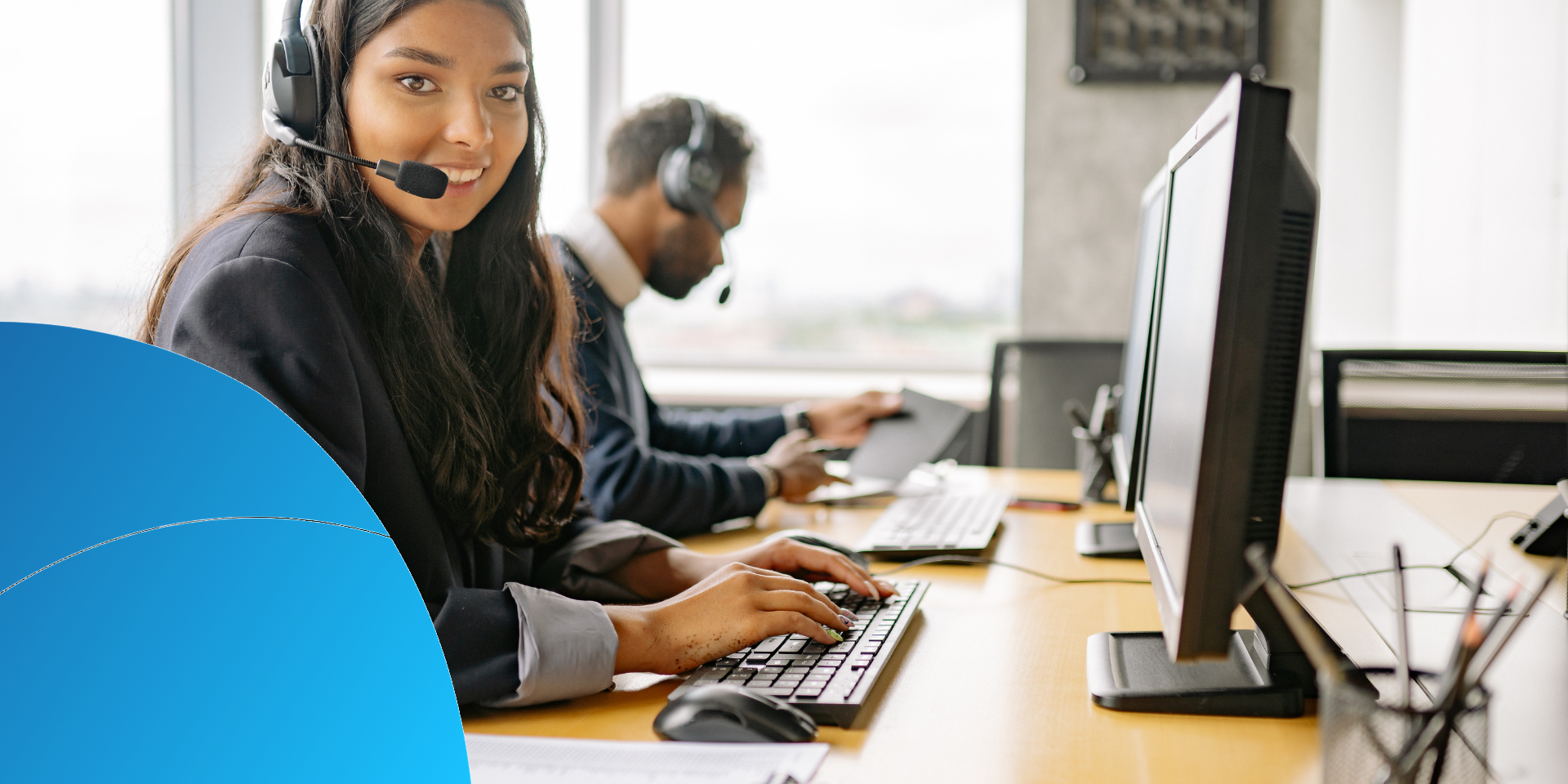 Kundenservice-Mitarbeiterin mit Headset am Arbeitsplatz, im Hintergrund ein Kollege