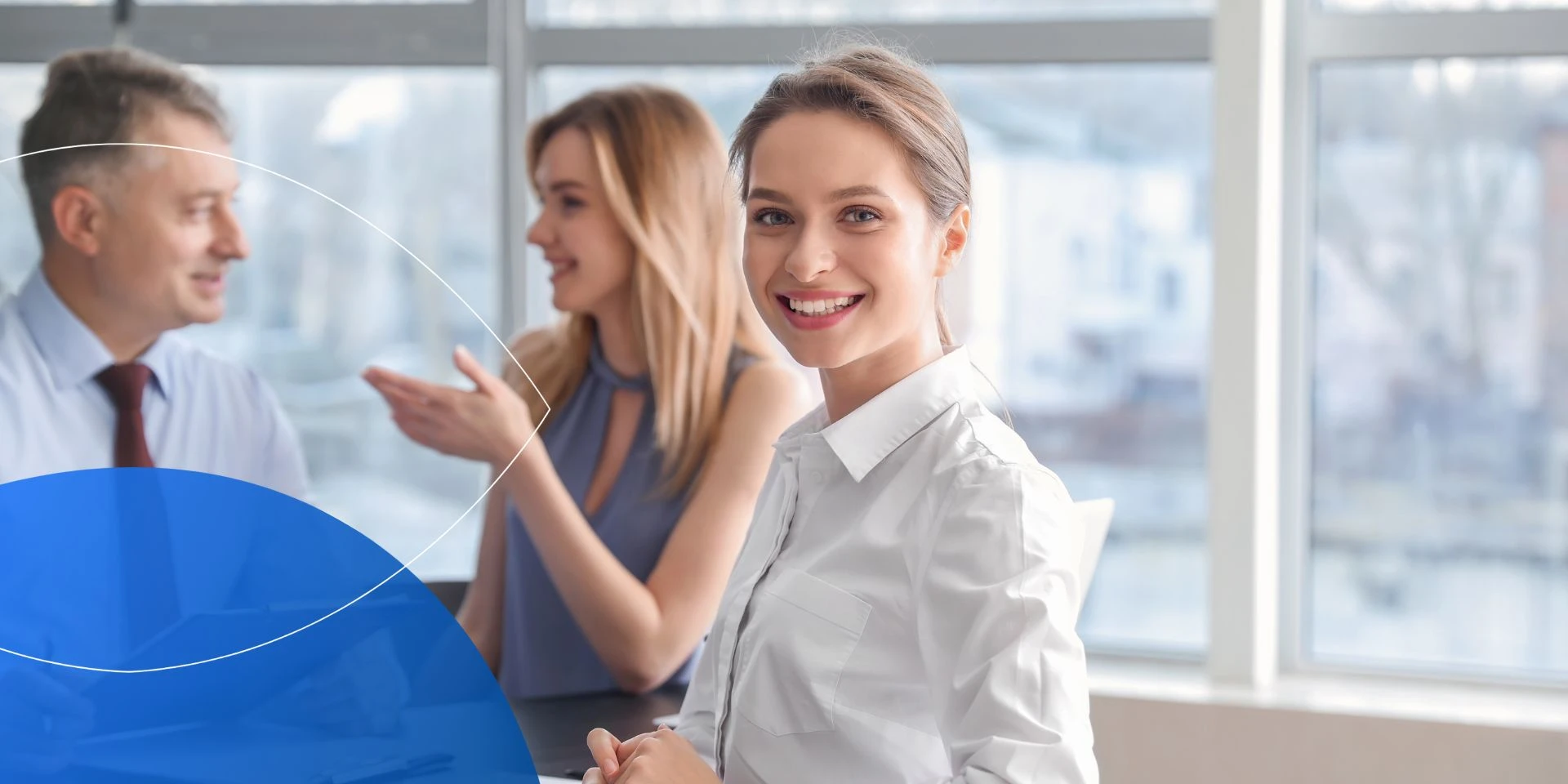 Woman sitting at desk and smiling to camera with another woman and man in the background 