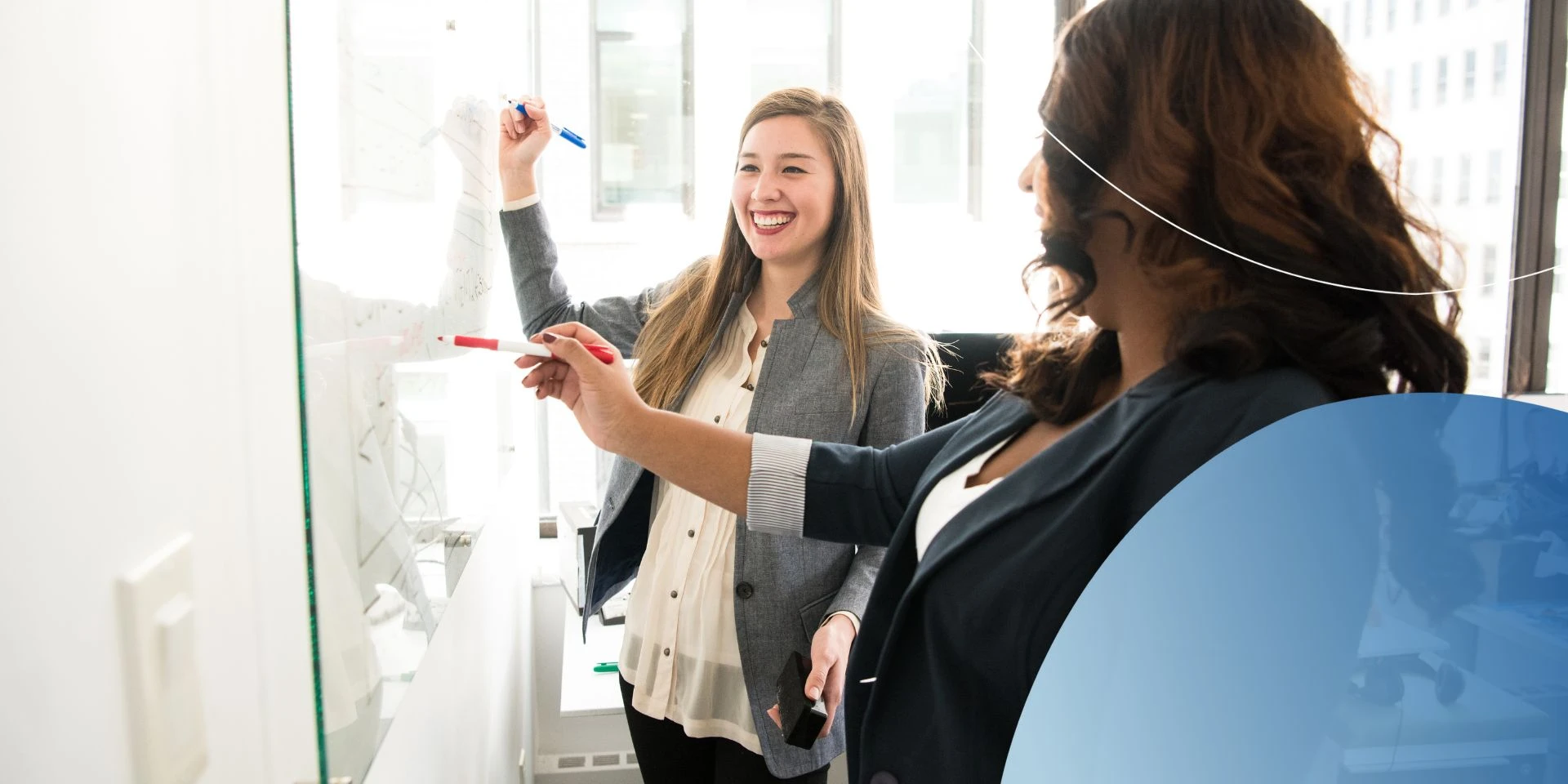 woman smiling and writing on white board next to another woman 