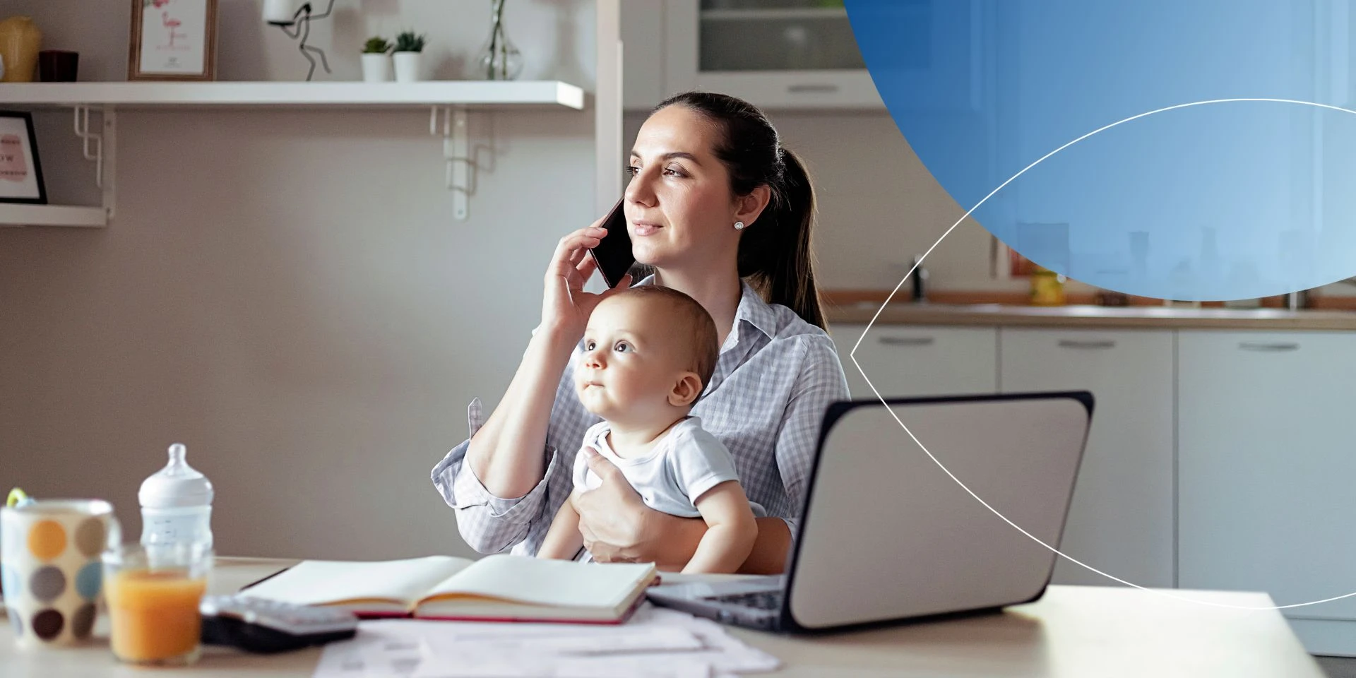 Woman sitting on a desk with laptop talking on the phone with a baby on her lap